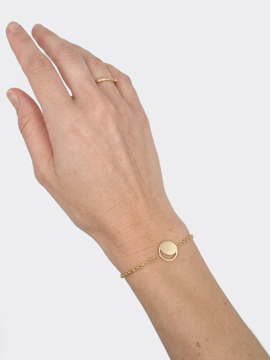 A delicate gold chain bracelet with a small flat circular disc moon phase charm, shown worn on a person's wrist alongside a matching gold ring on their finger, set against a plain light gray background.